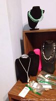 Five black velvet necklace display stands presenting various costume jewelry pieces including beaded necklaces and a silver-tone chain with pendant. Assorted jewelry in plastic bags is on the table.