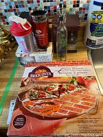 Photo showing boxed 13 inch Brick Oven pizza stone with wire rack, positioned on countertop with other kitchen items.