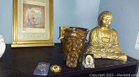 Gold Buddha statue, wood carved Jesus head, and small religious figurines on black surface. Framed artwork in background.