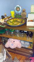 Shelf with Mexican decorative plate, blue and yellow tall container with floral smiling face decoration, wooden Holland boy figurine wearing textured clothes, coconut monkey figurine, shells in a white box, wooden paddle, various glass jars including peanut butter jars.