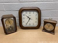 Photo of three mantle clocks with wooden frames on a table showing different styles and sizes including a large Bulova wall clock.