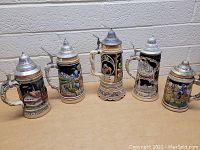 Group view of five German beer steins showing detailed colorful artwork and pewter lids.