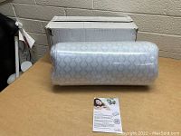 Wedge pillow wrapped in protective plastic packaging, patterned fabric visible, placed on a table with a box behind it.