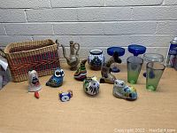 Woven basket, metal pitcher, blue and white ceramic vase, multiple animal-shaped ceramic figurines, and several pieces of glassware arranged on a table.