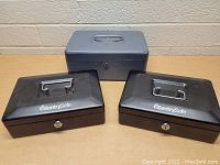 Photo showing three cash boxes: two black SentrySafe boxes and one grey metal box, placed on a beige surface with a brick wall background.