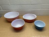 Four vintage Pyrex mixing bowls in orange and blue with white interiors arranged on wood surface against brick wall.