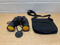 Black binoculars with yellow lenses and textured grips beside black fabric carrying case with shoulder strap and flap, displayed on wooden surface against brick wall.