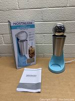 Photo of Nostalgia milkshake maker with its box and user manual against a concrete wall background.