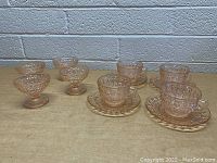 Photo showing 4 pink glass cups with handles placed on 4 matching saucers on a beige surface in front of a light-colored brick wall.