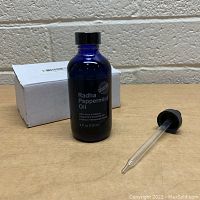 Blue glass bottle of Radha Beauty Peppermint Essential Oil 4oz with black screw top and glass dropper lying beside it, white box in background.