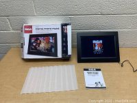 Photo showing the RCA digital photo frame powered on displaying the RCA logo, alongside the original box, user guide, and protective padding.