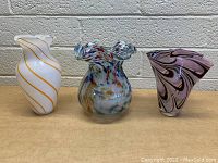 Frontal view of three blown glass vases: white with gold swirl on left, multicolored ruffled rim in center, purple with black swirl on right.