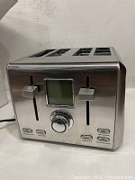 Side angle showing brushed stainless steel toaster with four slots, two vertical sliders, central dial, and buttons. Visible dent on side panel.
