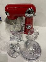 Cuisinart red stand mixer base with various wrapped accessories including dough hook, whisk, paddle, pour shield, and other small parts laid out on table.