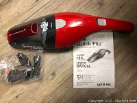 Dirt Devil Quick Flip handheld vacuum with red and black body, power button on handle, transparent dust container, user manual, charging adapter, and nozzle attachment on wooden floor.