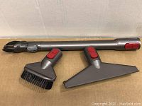 Three Dyson vacuum accessories arranged on cardboard surface including stiff bristle brush, wide nozzle mattress tool, and extendable wand with quick release buttons.