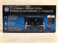 Box packaging of the HP 2.1 compact speaker system showing product image, branding, and features like 24W peak power, wired remote, and magnetically shielded drivers.
