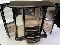 Open jewelry box showing the beige soft-lined compartment with 8-hook carousel, ring rolls, drawers, and mirrors on doors