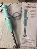 KitchenAid handheld immersion blender and clear plastic measuring cup beside original packaging box.