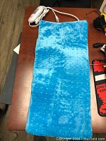 Blue rectangular heating pad with a plush micro-fleece textured surface and attached white power adaptor with control switch, laid flat on wooden surface.