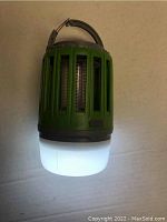 Green camping lantern and mosquito zapper placed on neutral flat background showing front with illuminated diffuser.