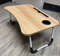 Photo showing the ergonomic laptop stand from a front angle on flooring, displaying foldable white metal legs with black rubberized areas and oak woodgrain top with tablet slot and cup holder.