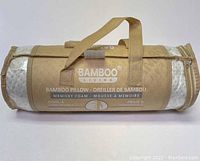Bamboo memory foam pillow in tan cylindrical carrying bag laying on white surface, showing front with branding and product description.