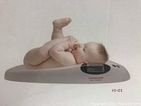 Baby scale shown with infant lying on it. White, curved design, digital LCD on side.