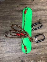 Top view of green plastic swing seat laid flat on wooden floor with attached brown rope featuring red adjusters and two removable black carabiners nearby.