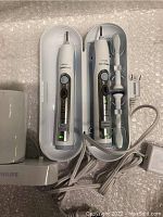 Two Philips Sonicare electric toothbrush handles in white hard travel cases, visible control buttons and LED displays. One brush head attached to right toothbrush. Power chargers and cords visible.