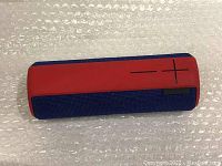 Red and blue UE portable Bluetooth speaker lying sideways on bubble wrap, showing top control symbols for volume plus and minus.