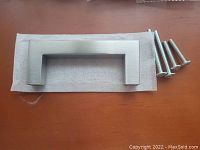 Single silver square cupboard handle with four included mounting screws placed on a towel on a table.