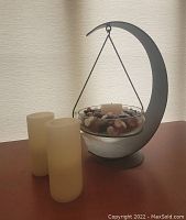 Hanging metal crescent shaped decor piece holding glass bowl with beads and a real candle inside.