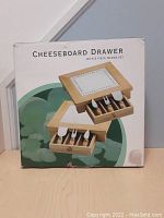 Box photo showing cheeseboard drawer with 3-piece wedge set of utensils on front of box.