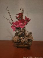 Photo of large artificial flower decor showing bright pink flowers, twigs, seed pods, and a rustic square base with metal feet on a wooden surface.