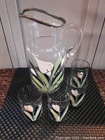 Clear glass pitcher with silver rim and hand painted white and pink flower with green leaves. Four matching glasses with same design placed around the pitcher.