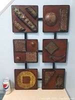 Metal wall art composed of eight square panels featuring various textured and embossed designs in rust and bronze colors, arranged vertically on a black frame. A standard soda can shown for scale.
