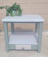 Side view of the newly refinished square side table showing whitewashed top and undershelf with sage green painted frame and legs, with a small drawer at the bottom center.