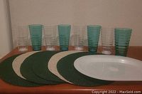 8 stacking outdoor plastic glasses, 4 teal and 4 clear, arranged in a row, with 6 round green and beige placemats in front of them on a wooden surface.