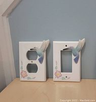 Photo showing two ceramic switch plates, one for a dual electrical outlet and one for a single light switch. Both have raised 3D hummingbirds with blue hues and floral accents on a white background.