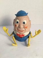 Front view showing Humpty Dumpty's face, blue hat, red bow tie, and yellow limbs.