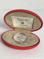 Silver coin displayed inside red velvet-lined presentation case with certificate inside lid.