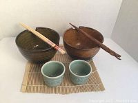 Set displayed on beige bamboo mat showing all items: 2 bowls, 2 cups, 2 pairs of chopsticks