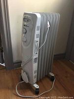 Side view of white Noma oil-filled radiator heater with multiple heating fins and caster wheels, plugged in with power cord visible.