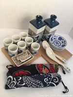 Two sake bottles, eight sake cups, four small dishes, two spoons, two rectangular trays, two fabric bags arranged on a wooden tray.