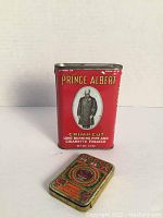 Front view of large red Prince Albert Crimp Cut tobacco tin with a portrait and smaller decorative tin beside it