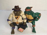 Photo showing two vintage TMNT action figures: Donatello with jacket, hat, and accessory, and Michelangelo with orange mask and bands, standing upright on white surface.