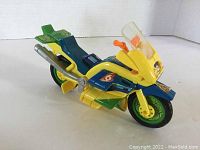 Side view of yellow, blue, green Playmates 1980s TMNT motorcycle toy with decals and exhaust detail.