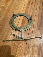 Brass ship porthole frame with mounting bolts and clasps laid flat on wooden floor next to the brass scale.
