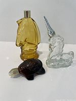 Three Avon animal-shaped perfume bottles on white surface: yellow horse head, clear unicorn with silver horn, and brown turtle.